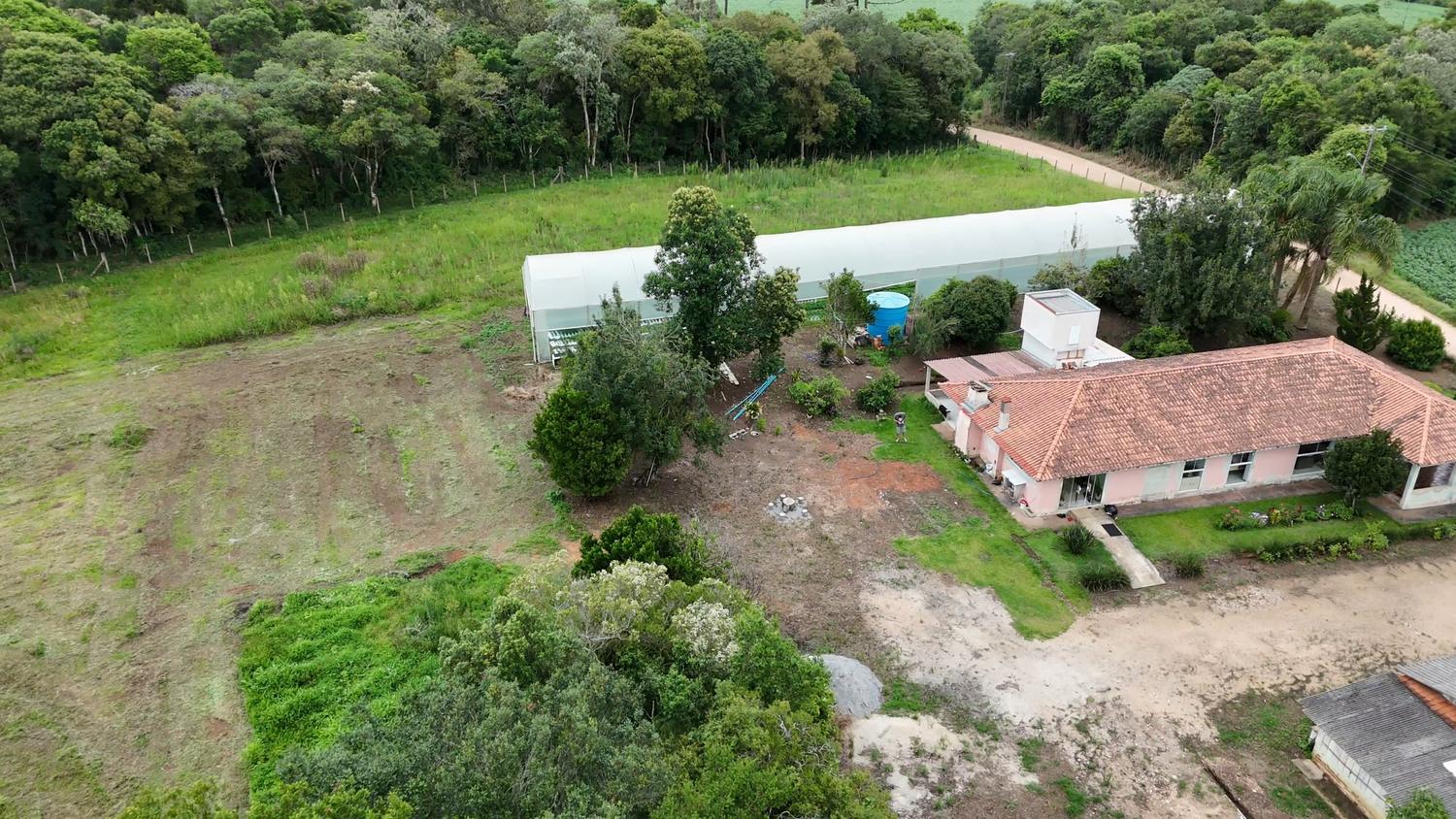 Vista aérea da casa sede: volume térreo alongado com telhado cerâmico e fachada em tom claro, estufa alongada logo atrás, caixa d'água azul, pátio de terra e ampla área plana à esquerda.